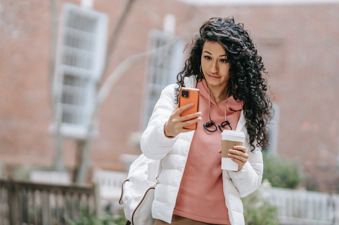young ethnic female drinking takeaway coffee and using smartphone on street