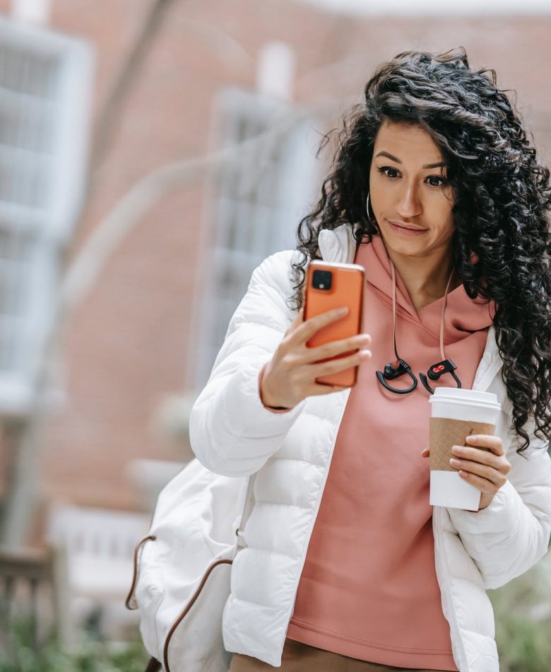 young ethnic female drinking takeaway coffee and using smartphone on street